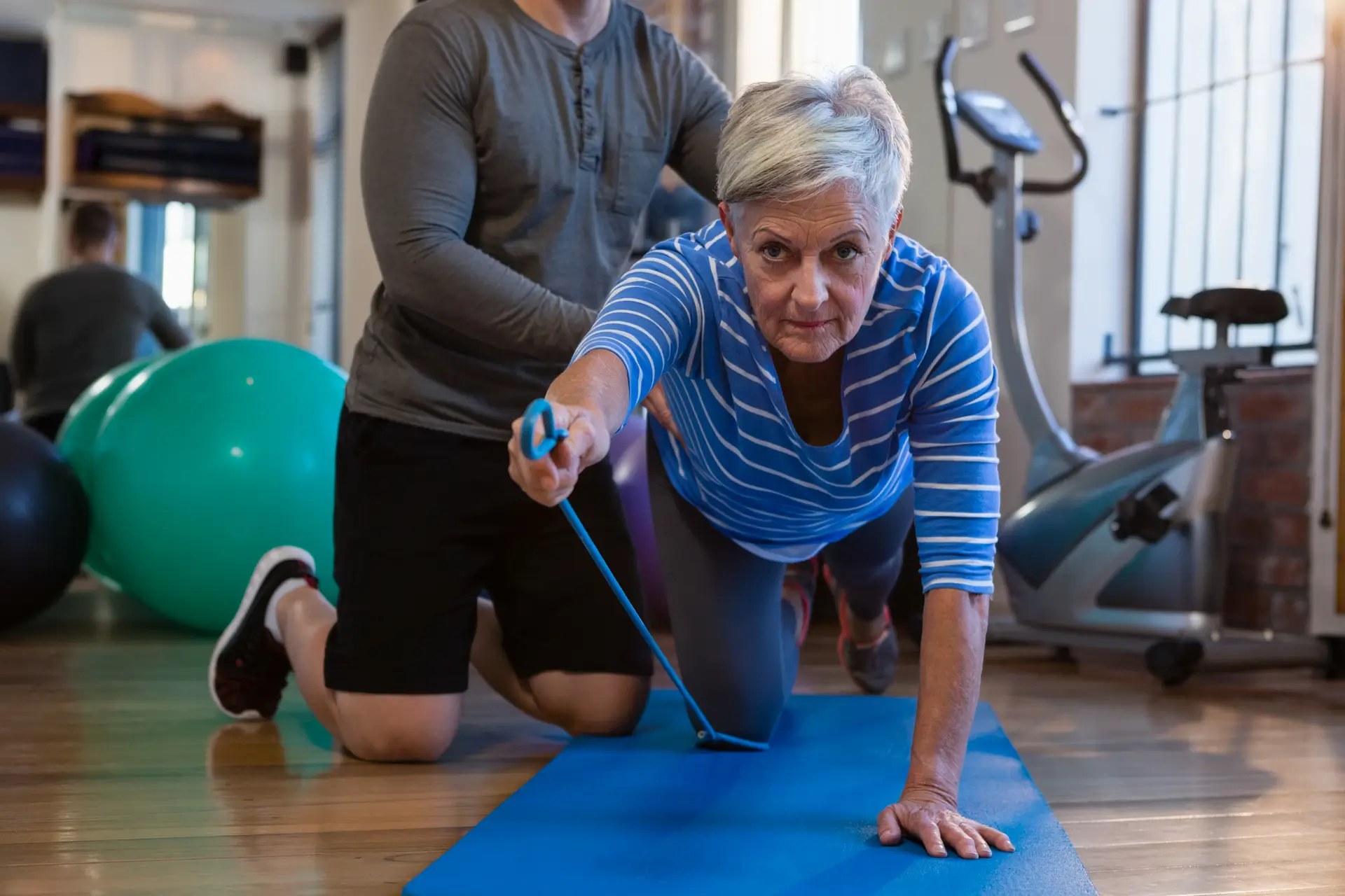 Elderly woman doing Therapeutic Exercises with a resistance band while a trainer assists her on a mat in a physiotherapy gym.