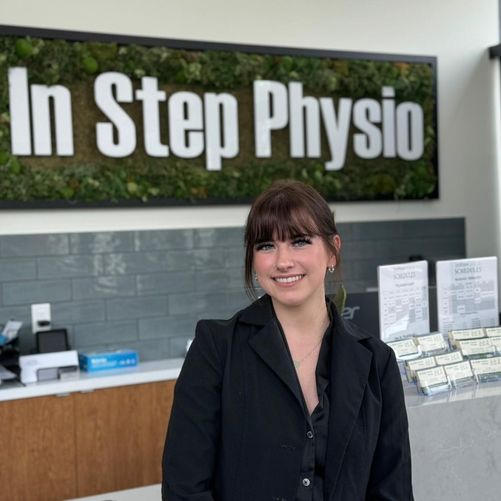 Mackenzie Carter - Provisional Psychologist In Instep Physical Therapy Smiling At Clinic Reception Desk With Branded Signage In Background.