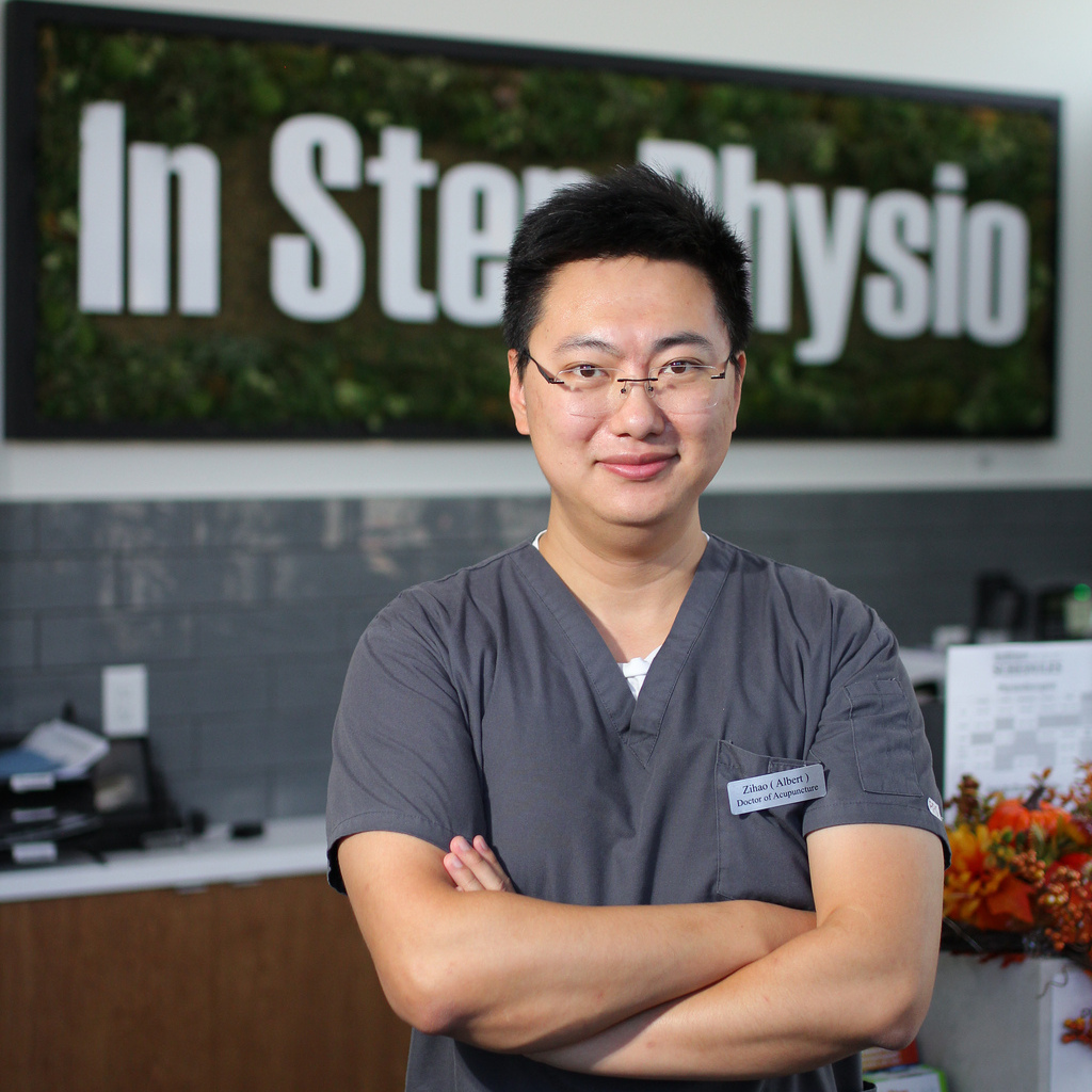 Zihao (Albert) Wang - Acupuncturist In Instep Physical Therapy Standing With Arms Crossed At Clinic Reception, Wearing Scrubs And Glasses.